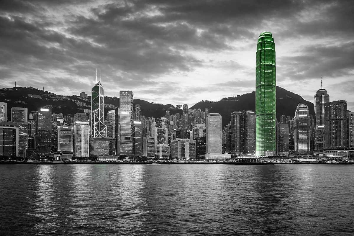 Black-and-white image of the Hong Kong skyline with the city’s skyscrapers selectively highlighted in green (#009650), reflected across Victoria Harbour under a dramatic cloudy sky.