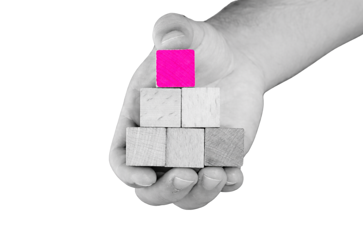 Black-and-white close-up of a hand holding stacked wooden blocks in a pyramid, with the top block highlighted in bright pink against a transparent background.