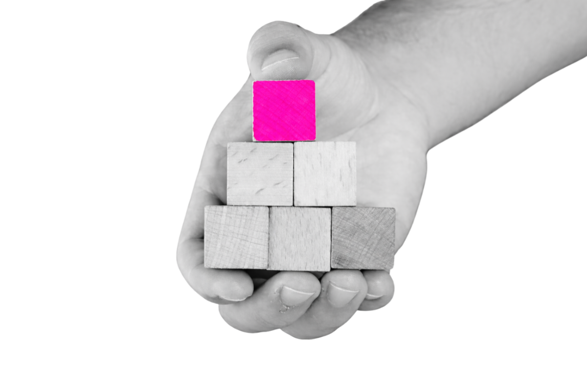 Black-and-white close-up of a hand holding stacked wooden blocks in a pyramid, with the top block highlighted in bright pink against a transparent background.